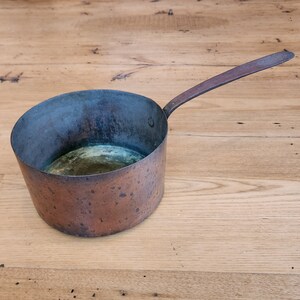 以下が含まれることがあります： 長い取っ手のある銅製の鍋。鍋は緑青がかっており、少し曇っている。