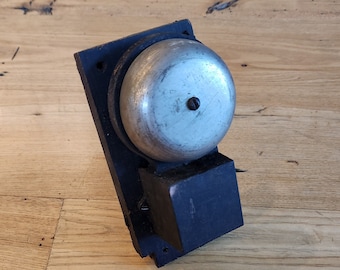 May include: A black metal doorbell with a round, silver bell. The bell is mounted on a wooden surface.