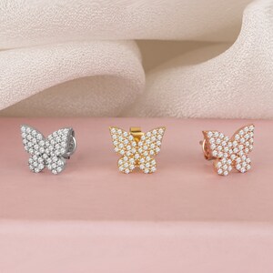 May include: Three butterfly-shaped earrings in silver, gold, and rose gold. Each earring is encrusted with small, sparkling stones. The earrings are displayed on a pink surface with a white fabric backdrop.