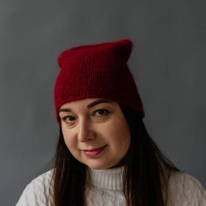 Puede incluir: Una mujer con un gorro de punto rojo y un suéter de punto blanco con cable. Está mirando a la cámara con una leve sonrisa.