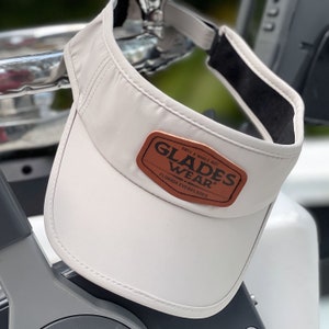 May include: A white visor with a brown leather patch that says "Glades Wear" and "Est'd a While Ago Florida Everglades".