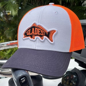 May include: White and orange trucker hat with a black and orange fish patch that says "GLADES" on it. The hat has a gray brim.