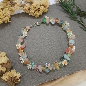 May include: A delicate necklace made with a variety of colorful stones and small flower charms. The necklace is made with a silver chain and features a mix of green, blue, pink, and orange stones.