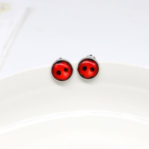 May include: A pair of round stud earrings featuring a vibrant red background with black circular spots, mimicking the appearance of a ladybird. Each earring is encased in a polished silver-toned metal setting with a smooth, glossy finish.
