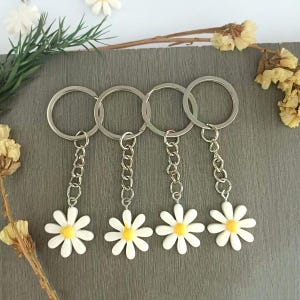 Op de afbeelding: Vier sleutelhangers met madeliefbloemen, zilverkleurige kettingen en sleutelringen. De madeliefhangers hebben witte bloemblaadjes en gele centra. De sleutelhangers worden gepresenteerd op een grijze ondergrond met decoratieve bloemen en groen.