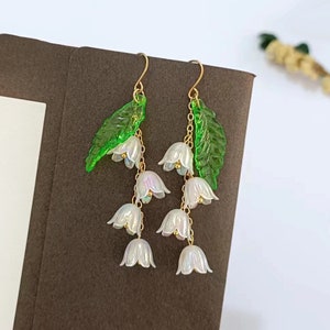 May include: A pair of dangle earrings featuring a cluster of white lily of the valley flowers and green leaves. The flowers are iridescent and have a pearlescent finish. The earrings are made with gold-toned chains and hooks.