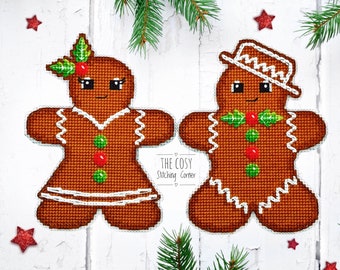 May include: Two gingerbread people cross-stitched patterns. The gingerbread person on the left is a female with a green holly sprig on her head. The gingerbread person on the right is a male with a brown hat. Both gingerbread people have green and red buttons and white icing.