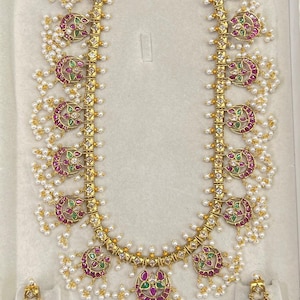 May include: A gold-toned long necklace and matching pair of earrings featuring crescent-shaped motifs inlaid with small red and green stones, accented with clear white stones and dangling clusters of tiny white seed pearls throughout the design.