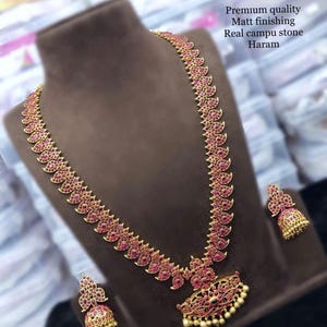May include: A gold-toned necklace and matching earrings set. The necklace features a long, ornate design with red gemstones and a pendant. The earrings have a similar design. The text in the image says "Premium quality, Matt finishing, Real campu stone, Haram."