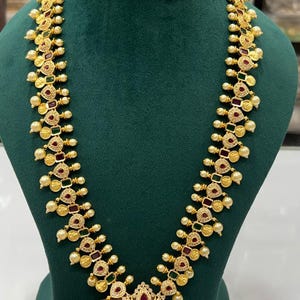May include: A gold necklace and earring set with detailed patterns. The necklace has a central pendant with a figure, red and green accents, and pearl-like beads. The earrings match the necklace design, forming a complete jewellery set.