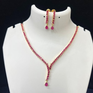 May include: A gold-toned jewelry set featuring a necklace and matching earrings. The necklace and earrings are adorned with vibrant, oval-shaped, red gemstones. The necklace has a Y-shaped pendant with a teardrop-shaped gemstone at the end. The earrings are a linear design.