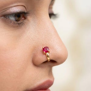 May include: A gold nose ring featuring a square, red gemstone. The ring is worn on a person's nostril. The gemstone is a vivid red, and the gold band has a smooth, polished finish.