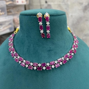 May include: A jewelry set consisting of a double-strand necklace and matching drop earrings. The pieces feature rectangular, deep pink stones alternating with small, round, clear white stones set in a silver-toned metal frame.