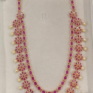 May include: A gold necklace and earring set. The necklace has a double strand design with oval ruby-coloured stones and floral details. Small pearl-like beads hang from the floral elements. The earrings match the necklace's design.