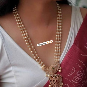 May include: A three-strand pearl necklace with a gold pendant featuring a floral design and green stones. The pendant is attached to a chain with a pearl drop. The necklace is worn by a person with a white shirt and a red saree. The text "Hot selling 🔥🔥🔥" is visible in the image.