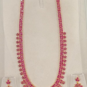 May include: A gold necklace and matching earrings set, featuring pink gemstones. The necklace has a long, symmetrical design with a curved base. The earrings have a floral design. The set is presented on a white background.