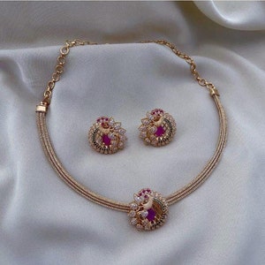 Puede incluir: Un collar de oro con un colgante de diseño de pavo real y pendientes a juego. El colgante y los pendientes presentan piedras rosas y verdes.