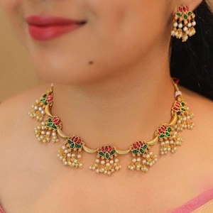 May include: Gold-toned necklace and earring set. The jewelry features floral designs with red and green accents, and small white pearl-like beads. The necklace and earrings are worn on a person.