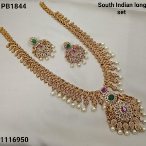 May include: A gold-toned South Indian long set, including a necklace and earrings. The necklace features a pendant with green and purple stones, surrounded by white gems and dangling pearls. The earrings have a similar design. The set is labeled "PB1844" and "1116950".
