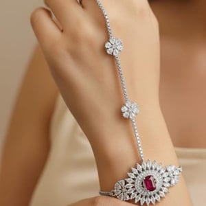 May include: A silver hand chain featuring a ring and bracelet connected by a delicate chain. The ring and bracelet have floral designs with clear stones. The bracelet has a central red stone.