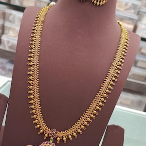 Op de afbeelding: Een goudkleurige ketting en bijpassende oorbellen set. De ketting heeft een ingewikkeld ontwerp met kleine, ronde accenten en een hanger met rode en groene edelstenen. De oorbellen hebben een vergelijkbaar ontwerp. De sieraden worden getoond op een bruine mannequin.