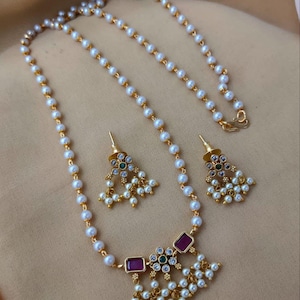 May include: A gold-toned necklace and matching earring set featuring white pearls strung along a delicate chain. The necklace pendant and earrings are adorned with rectangular red stones, small green center stones, clear crystals, and dangling pearl accents.