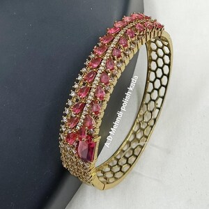 May include: A gold-toned hinged bangle bracelet featuring a central row of pear-shaped pink stones flanked by clear crystals. The side of the band displays a geometric honeycomb cutout pattern. The text A.D Mehndi polish kada is visible.