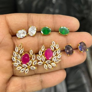 May include: A collection of stud earrings featuring two oval clear stones, two oval green stones, two oval dark purple stones, and two larger floral-inspired earrings with a central oval pink stone surrounded by marquise-cut clear stones.