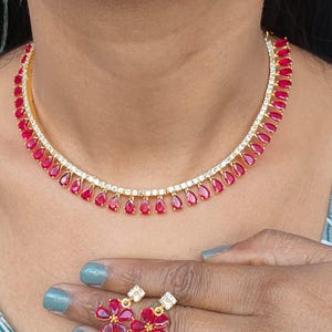 May include: A gold-toned necklace and matching earrings set. The necklace features a row of red teardrop-shaped stones and clear crystals. The earrings are flower-shaped with red petals and a small square crystal at the center. The jewelry is worn on a person.
