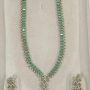 May include: A necklace and earring set with light green, rectangular stones set in silver-coloured metal. The necklace has a pendant with a teardrop-shaped green stone. The earrings have a similar design. The set is displayed on a light grey surface.