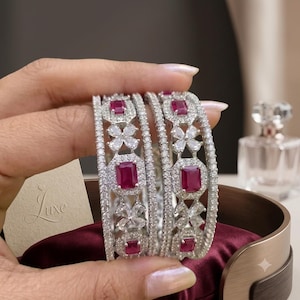 Peut inclure: Deux bracelets rigides argentés ornés d'un motif répétitif de pierres rouges rectangulaires et de pierres transparentes en forme de fleur. Les bracelets sont bordés de fines rangées de petites pierres transparentes étincelantes.