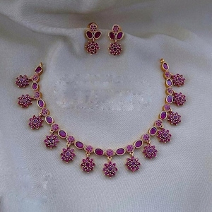 May include: A gold necklace with a pink stone design. The necklace has a chain with oval-shaped pink stones and dangling pink flower-shaped stones. The necklace comes with a matching pair of earrings.