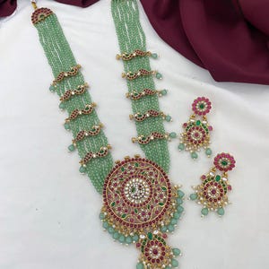 Pode incluir: Um conjunto de colar longo e ornamentado e brincos a condizer. O colar apresenta fios de contas verde claro, detalhes dourados e um pendente circular grande com detalhes vermelhos e verdes. Os brincos espelham o design do pendente. O texto "Long haram" é visível.