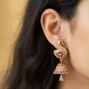 May include: A gold-toned dangle earring featuring a swan-shaped stud encrusted with small white and pink stones. A bell-shaped jhumka pendant hangs from the swan, decorated with matching pink and white stones and a single clear teardrop charm.