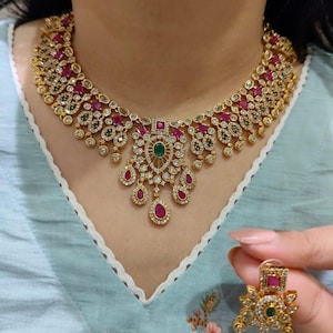 Puede incluir: Un conjunto de collar y pendientes dorados con piedras preciosas de color rojo rubí y verde esmeralda. El collar presenta un diseño detallado con un colgante central y pendientes a juego, mostrando un estilo de joyería clásico.