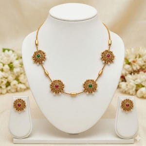 Puede incluir: Un collar de tono dorado con cuatro medallones circulares ornamentados, con piedras centrales alternas en verde y rosa, acentuados por pequeñas perlas blancas. El conjunto incluye dos pendientes circulares a juego con piedras rosas.