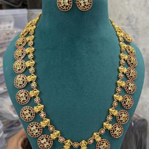 Op de afbeelding: Een goudkleurige ketting en bijpassende oorbellen set. De ketting heeft cirkelvormige hangers met rode, witte en groene edelstenen. De oorbellen hebben een vergelijkbaar ontwerp. De sieraden worden getoond op een groene mannequin.