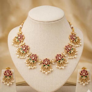 Puede incluir: Un collar dorado y un conjunto de pendientes a juego. El collar presenta colgantes con forma de flor con detalles rojos y verdes, adornados con pequeñas perlas blancas. Los pendientes reflejan el diseño del collar. Las joyas se exhiben en un maniquí.