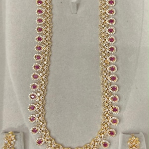 May include: A gold-toned necklace and matching earring set featuring teardrop-shaped pink stones surrounded by small clear white crystals. The necklace has a long, U-shaped design with repeating floral-inspired links and a central pendant.