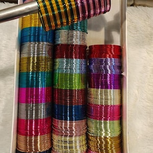 May include: A collection of thin, metallic bangles in a wide variety of vibrant colors including red, blue, green, gold, silver, pink, and purple. The bangles are organized in stacks within a white box, with some held on a metal rod.