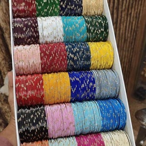 May include: A box filled with colorful bangles. The bangles are arranged in rows, showcasing a variety of colors including red, green, navy, gold, maroon, white, turquoise, yellow, silver, blue, black, pink, orange, and magenta. Each bangle is adorned with a subtle, glittery pattern.