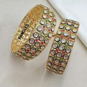 Peut inclure: Deux bracelets dorés ornés de pierres précieuses claires, rouges et vertes. Les bracelets sont décorés de nombreux pierres rondes. Le mot "BANGLES" est imprimé sur l'un des bracelets. L'image comprend également le texte "PB6019", "2/4 2/6 2/8" et "1114000".