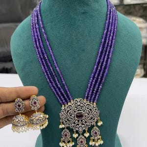 Op de afbeelding: Een paarse kralenketting met een grote, sierlijke hanger met een centrale paarse steen en heldere strass steentjes. De hanger is bevestigd aan een ketting met parel- en paarse kralenaccenten. Passende oorbellen met een vergelijkbaar ontwerp zijn ook inbegrepen.