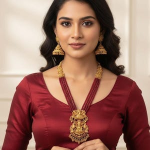 May include: A woman wearing a gold necklace with a pendant and matching earrings. The necklace has a multi-strand design with red beads and a detailed pendant. The earrings are bell-shaped. The woman is wearing a maroon blouse.