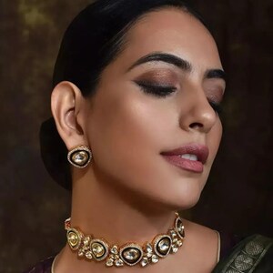 May include: A gold necklace and matching stud earrings. The jewellery set features oval-shaped, clear gemstones set in a gold frame with black detailing. The jewellery is worn by a person with dark hair and makeup.
