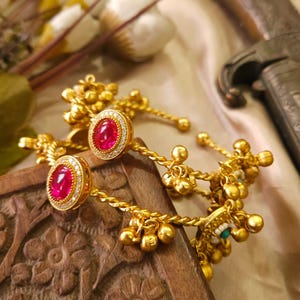 May include: Gold-toned jewelry with red gemstone accents and small dangling bells. The jewelry is displayed on a carved wooden surface, with a soft, neutral-toned background. The design features intricate details and a classic aesthetic.