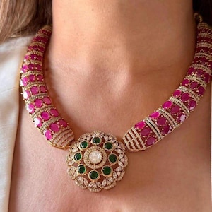 Puede incluir: Un collar dorado con un colgante central con una piedra grande y transparente rodeada de piedras verdes y piedras transparentes más pequeñas. La banda del collar está adornada con piedras rosas y detalles dorados, creando un aspecto lujoso.