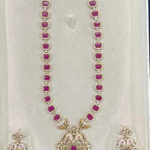 May include: A gold-toned necklace and matching earring set featuring rectangular, deep pink stones surrounded by small white crystals. The necklace has a large, ornate central pendant with dangling pink stones and white pearl-like accents.