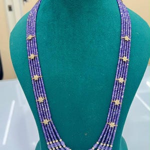 May include: A multi-strand necklace featuring five rows of faceted purple beads. Each strand is accented with gold-toned star-shaped spacers. The bottom center of the necklace features a single white pearl integrated into each of the five purple bead strands.