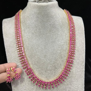 Op de afbeelding: Een gouden ketting en oorbellen set met roze edelstenen. De ketting is lang en heeft een aflopend ontwerp waarbij de edelstenen naar beneden toe groter worden. De oorbellen zijn klein en hebben een vergelijkbaar ontwerp als de ketting.